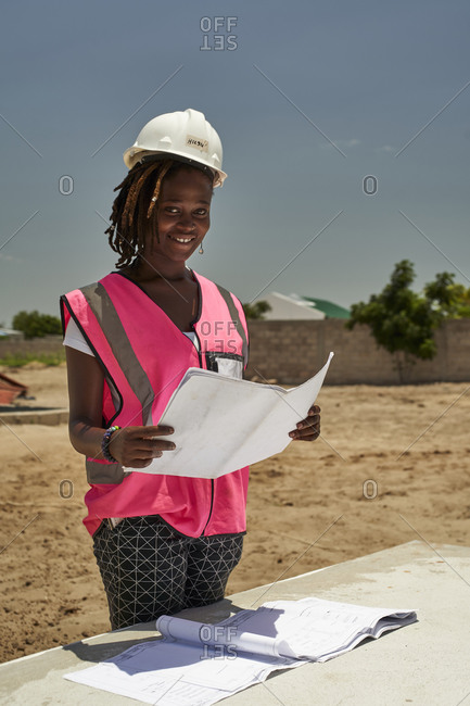 Smiling female architect holding blueprint while standing against clear sky at site during sunny day