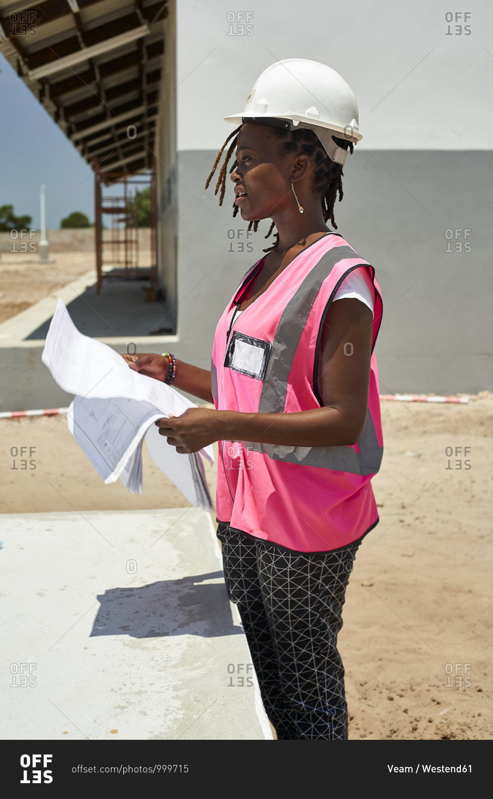 Female architect holding blueprint looking away while standing against school building at site
