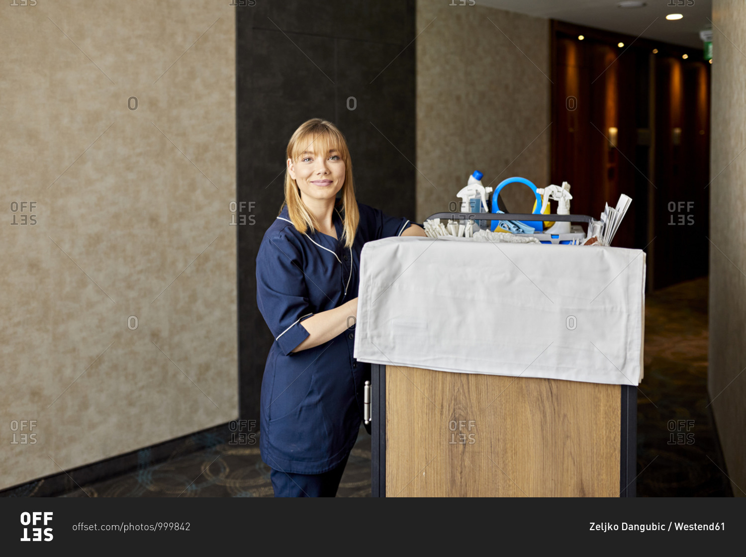 Smiling chambermaid with push cart standing in hotel corridor against wall