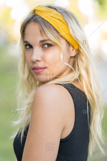 Side view of pretty young female with stylish yellow headband on blond woman looking at camera over shoulder on summer day in park