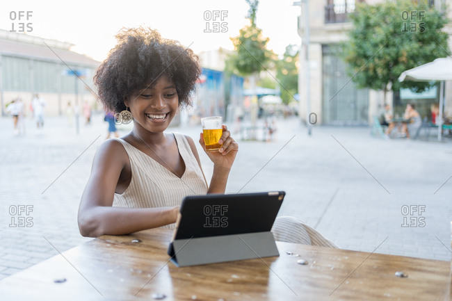 A smiling African-American woman sitting on the terrace, drinking a beer, consulting the tablet computer