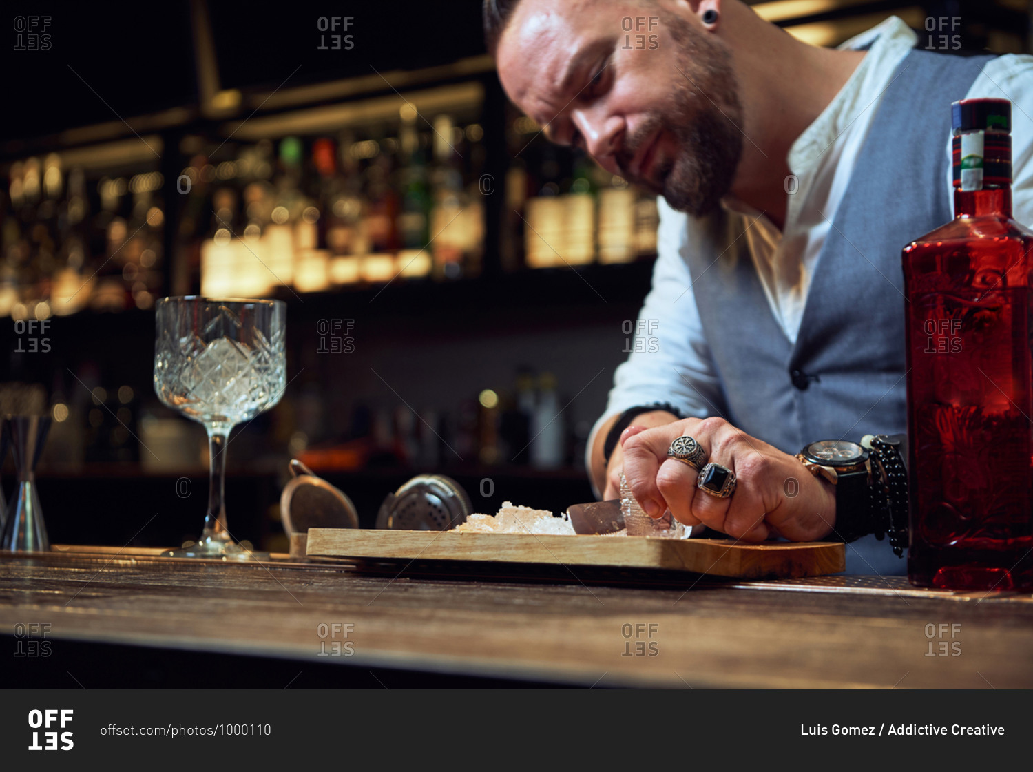 Professional bartender cutting ice cube with knife on wooden board while preparing a cocktail in the bar