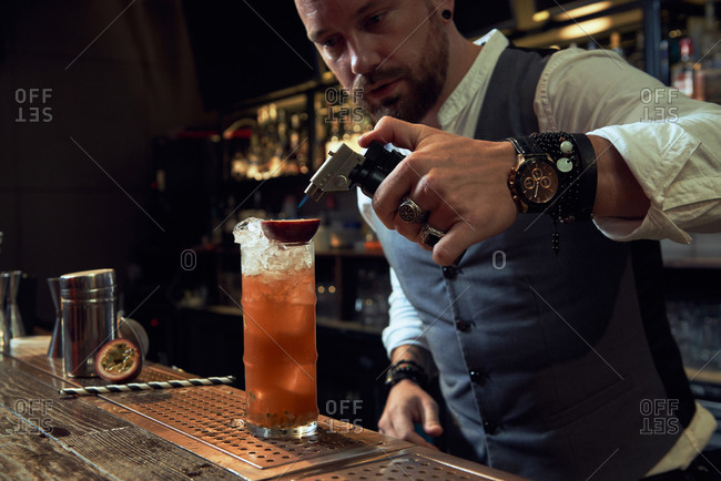 Professional young bartender using blowtorch to burn passion fruit in a glass with orange cocktail in a bar