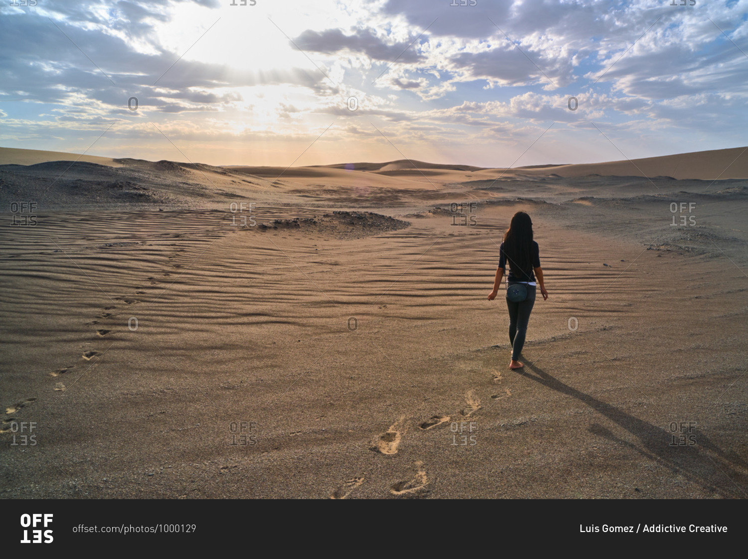 Back view of anonymous female tourist strolling in sandy terrain in desert with dry wavy surface under cloudy sky