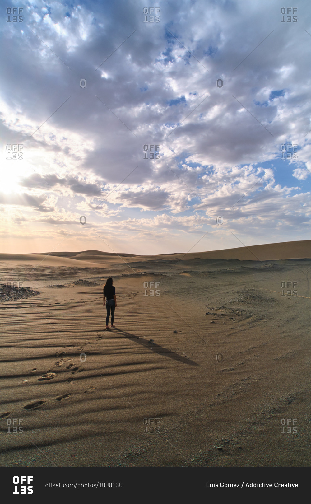 Anonymous female tourist strolling in sandy terrain in desert with dry wavy surface under cloudy sky at bright sundown