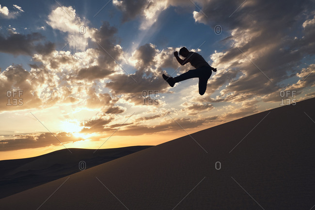 Silhouette of anonymous fit male dancer performing trick with reached leg above sandy terrain in desert at bright sundown at night