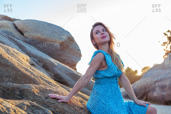 Low angle of woman traveler in summer dress posing on rough rock near sea during summer holidays