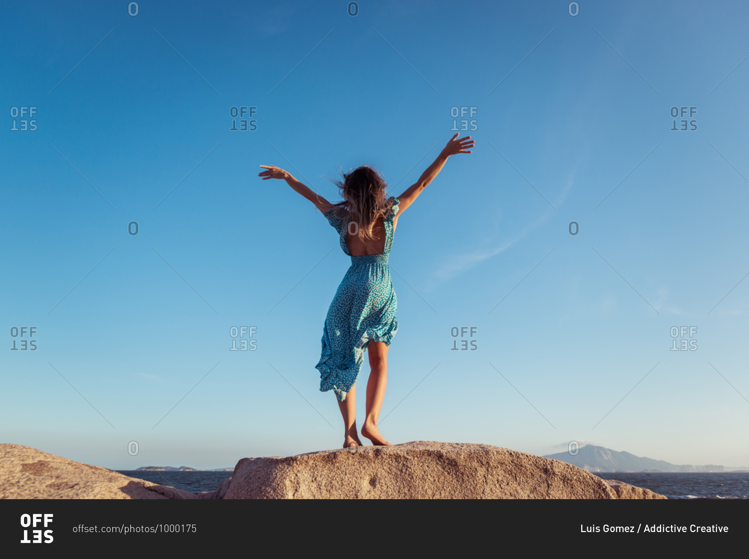 Back view of anonymous slim female in stylish light dress outstretching arms on rocky cliff against cloudless blue sky during summer holidays