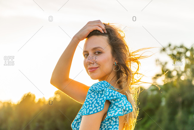 Optimistic young female looking at camera and touching hair on windy summer day during romantic sunset