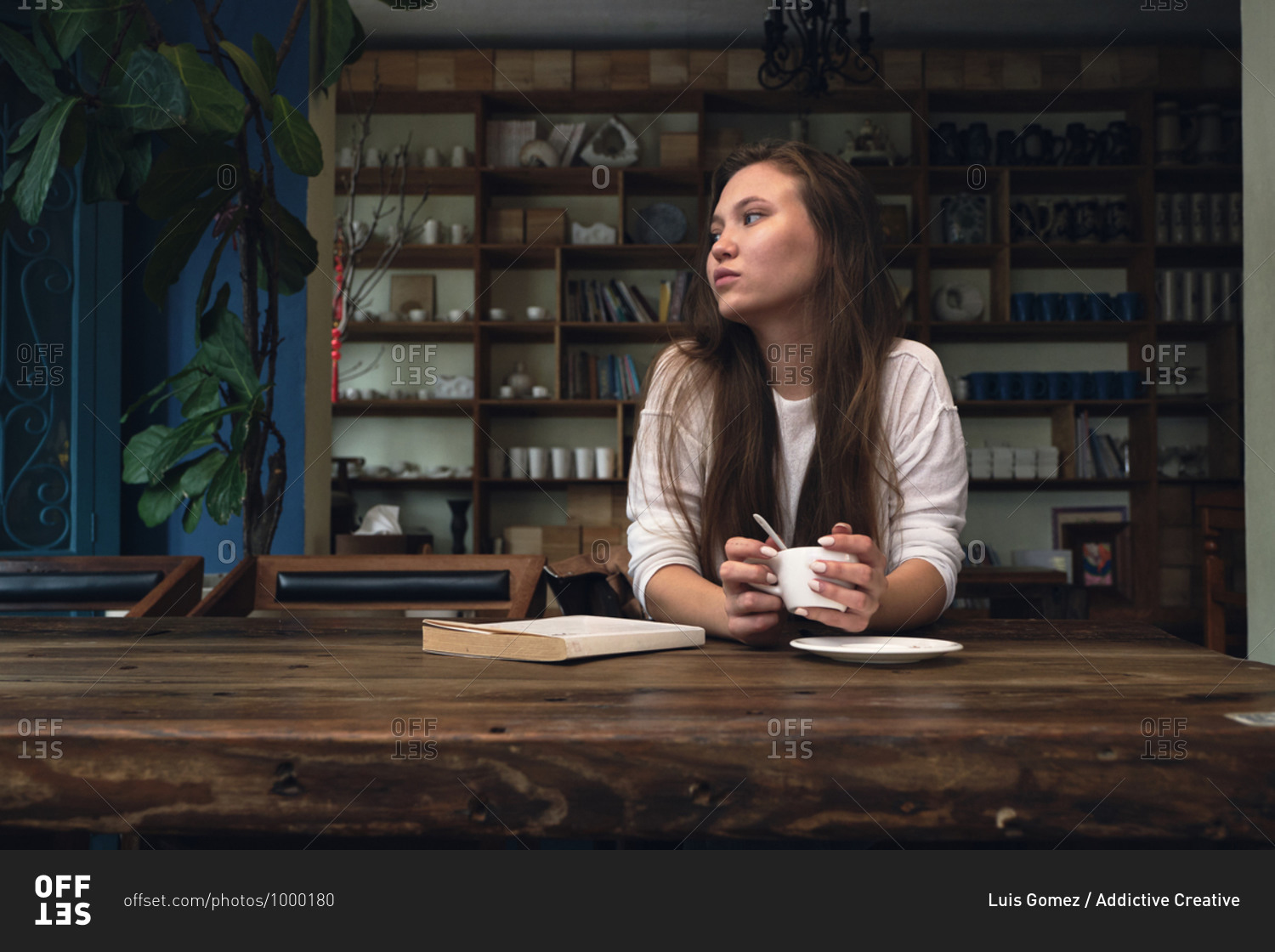 Pensive young woman with long hair relaxing at wooden table with book and drink coffee in cozy cafe