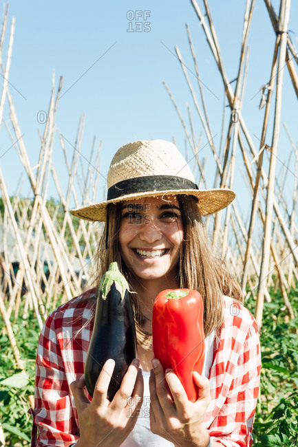 Content female horticulturist in straw hat and checkered shirt holding ripe red pepper and eggplant while standing near tomato bushes and wooden sticks under blue sky and looking at camera