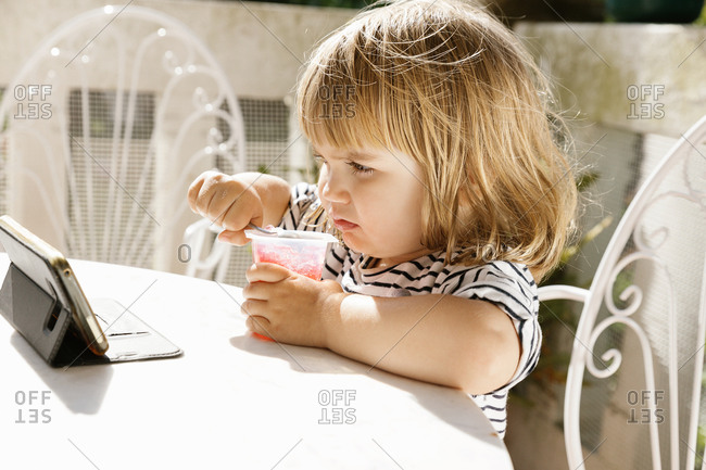 Cute little girl sitting at table in backyard and watching cartoon while enjoying delicious jelly dessert in summer