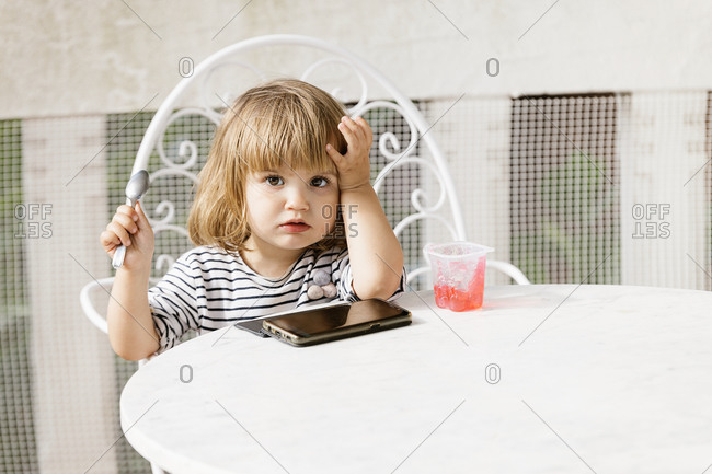 Cute little girl sitting at table in backyard and watching cartoon while enjoying delicious jelly dessert in summer looking at camera