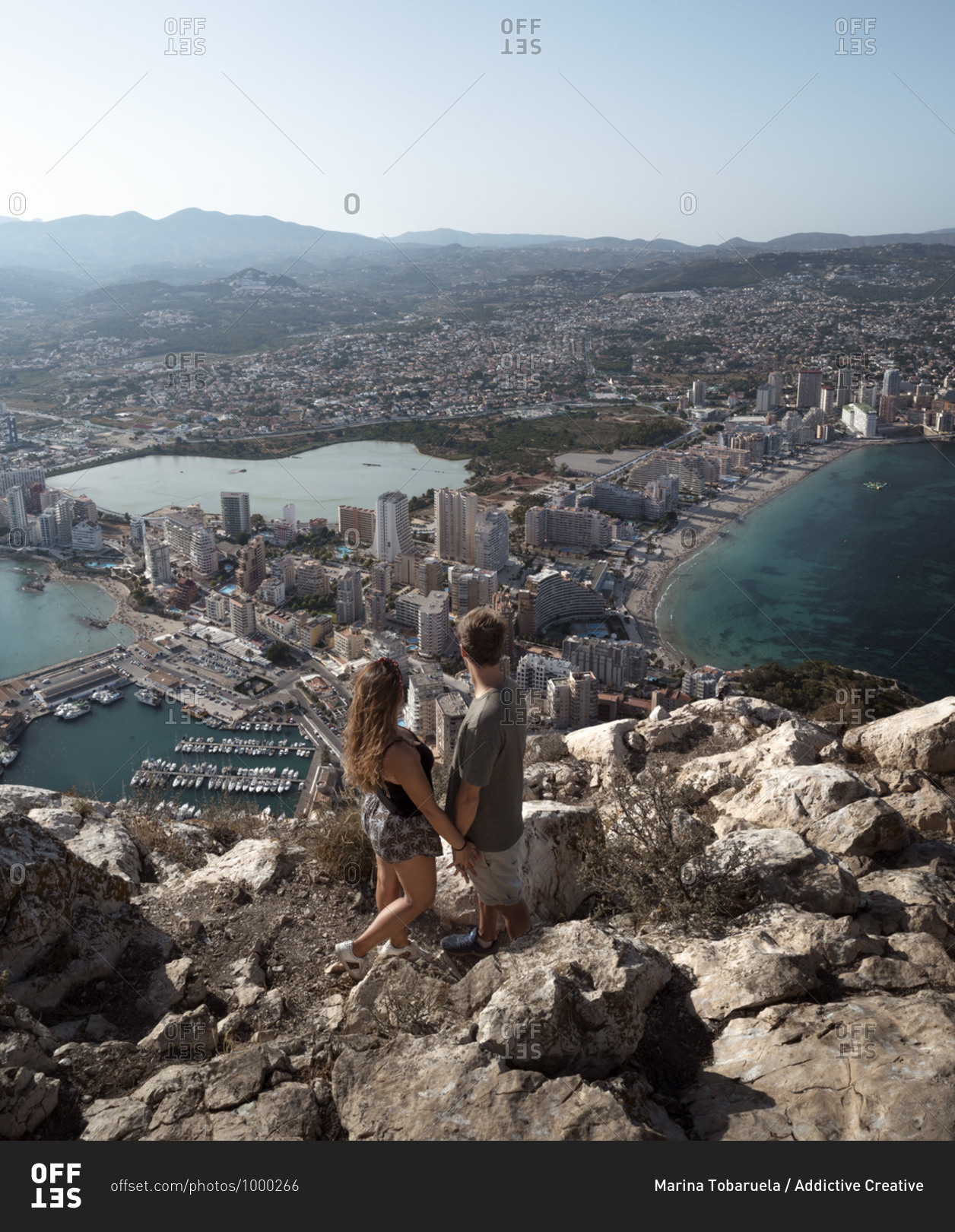 From above back view full body unrecognizable couple holding hands and observing fantastic view of Alicante while standing on high rocky hill viewpoint during sunny summer day