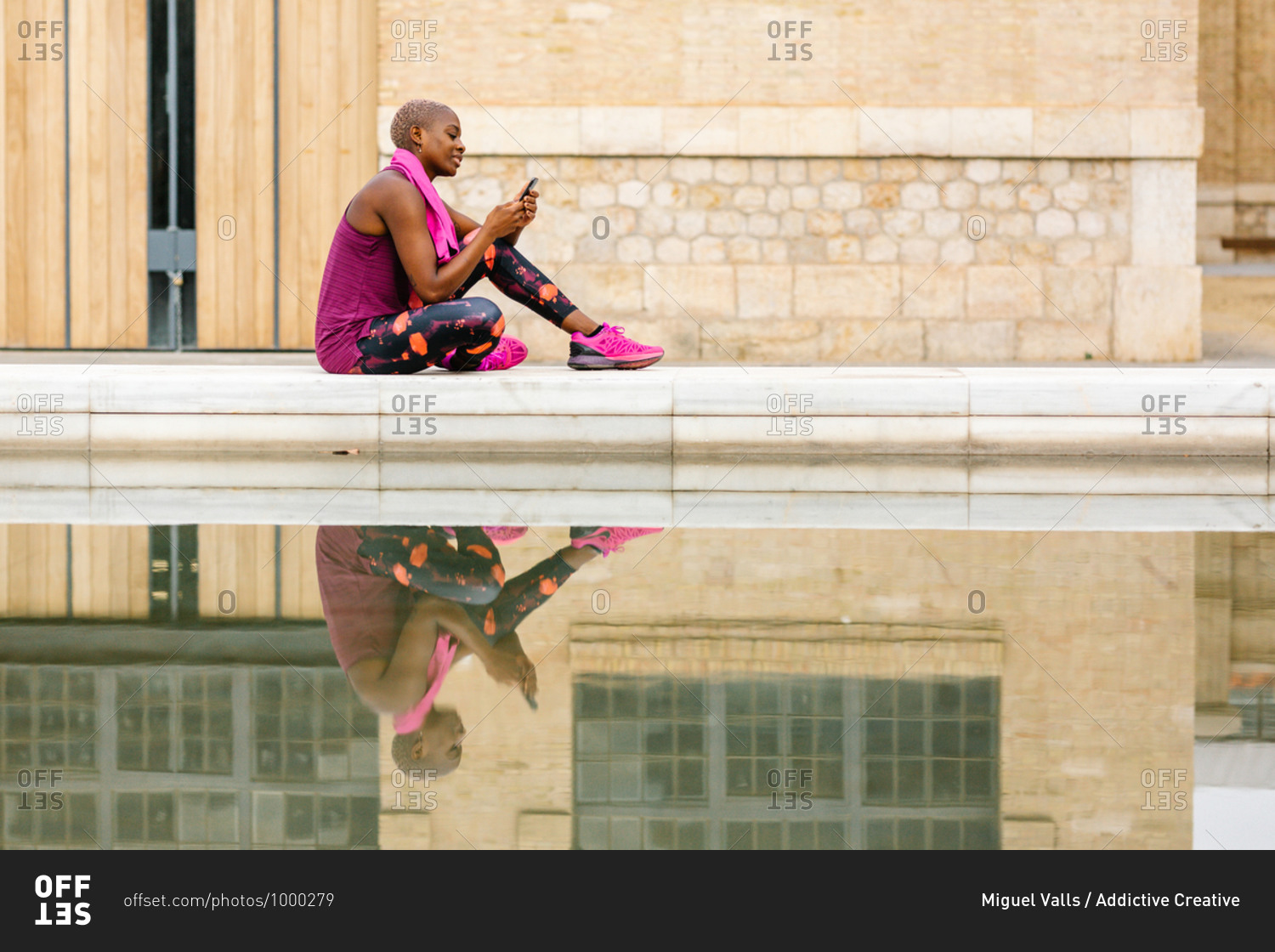 Side view of cheerful African American sportswoman in active wear with pink towel on shoulders using social media on cellphone while sitting on embankment near fountain and building in town
