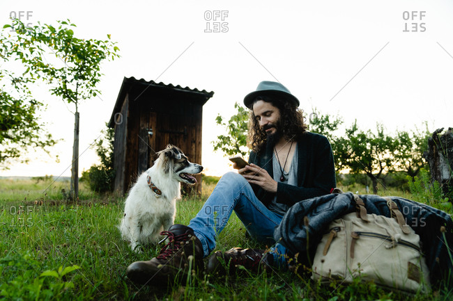 Delighted male hipster sitting on green filed in summer next to a dog and chatting on social media during weekend