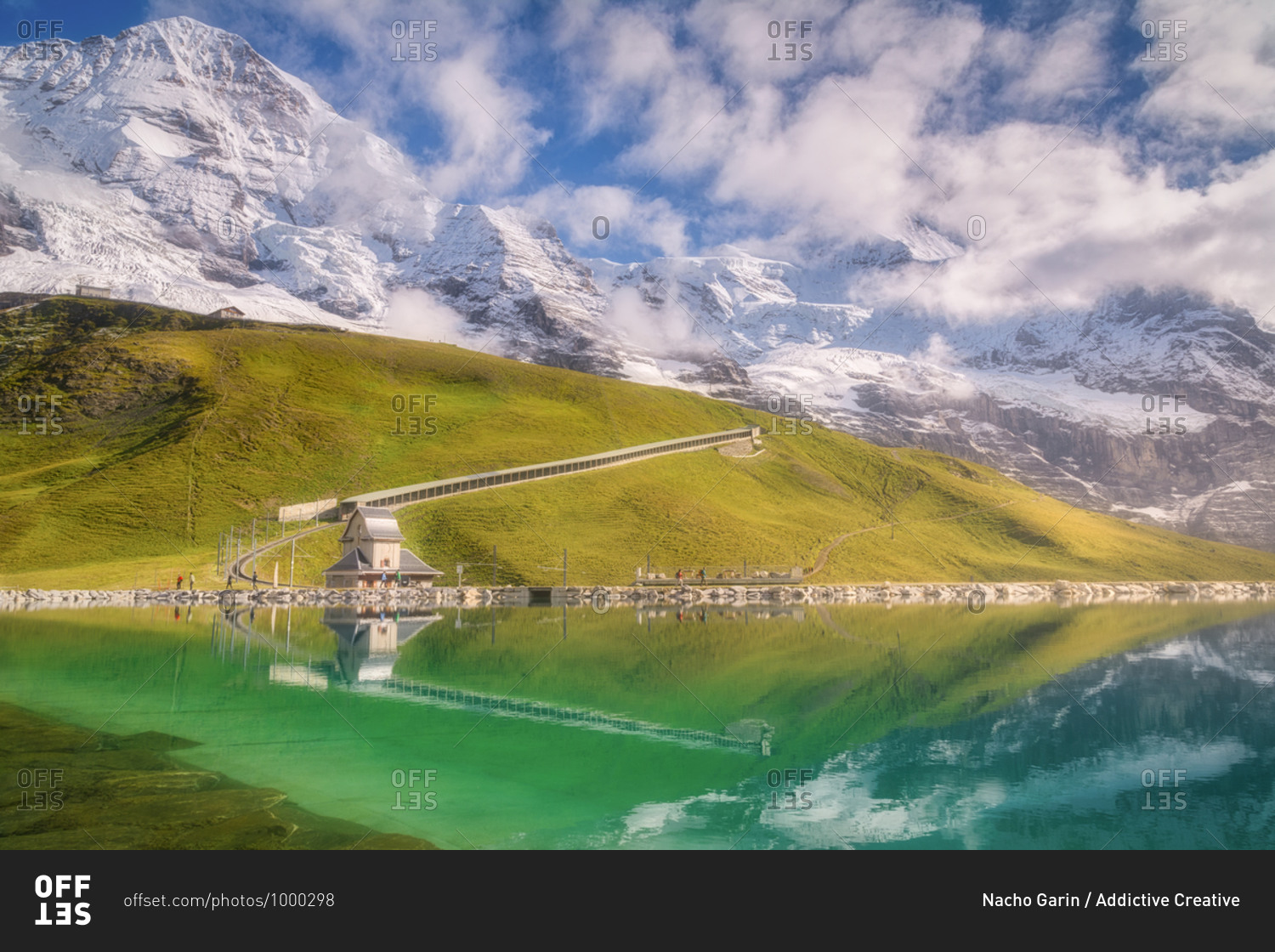 Majestic view of river with transparent water near green mountains with railroad and small house under snowy ridge with cloudy sky