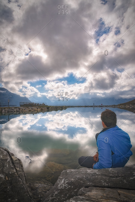 Back view of anonymous male tourist admiring magnificent lake with pure water surrounded by ridge and reflecting sky with clouds in daylight