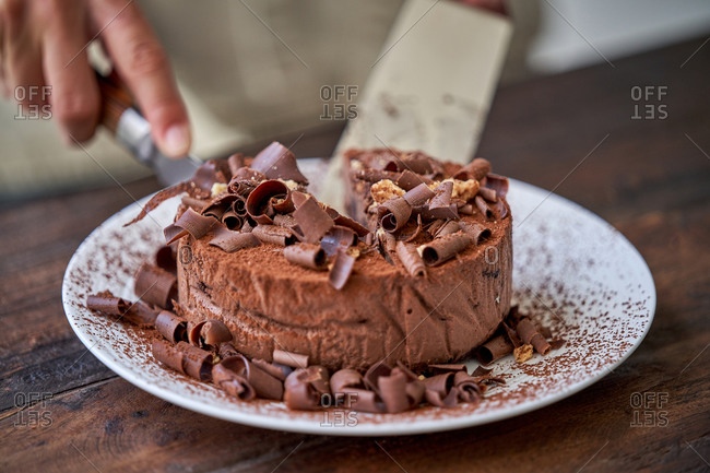 Crop of anonymous woman cutting chocolate cheesecake sprinkled with cocoa powder and decorated with chocolate shavings served on white plate