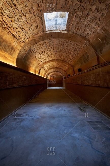 Long hallway with blots on floor under stone wall with rough uneven surface and arched entrance illuminated by artificial light