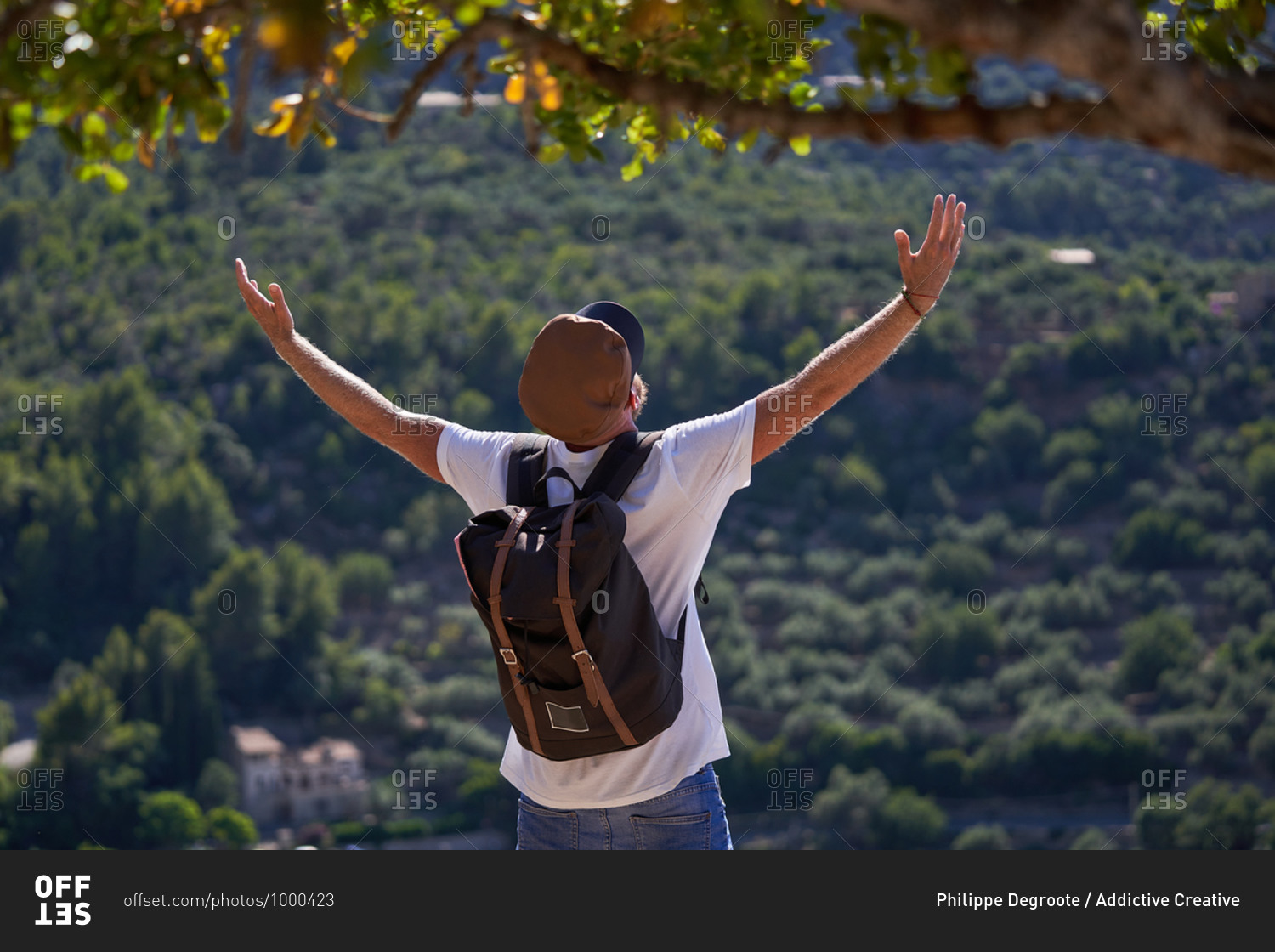 Back view of unrecognizable male hiker in cap with backpack standing with outstretched hands on hill against green forest and enjoying freedom during summer adventure