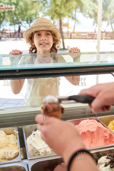 Crop ice cream shop assistant putting chocolate and vanilla ball in waffle cone for enthusiastic boy in straw hat