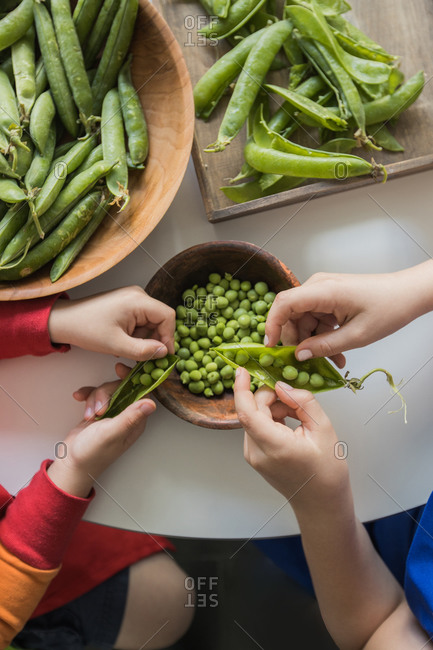 From above of crop unrecognizable boy peeling fresh green crunchy pea pods while sitting near sister in apron at table with wooden bowls in flat