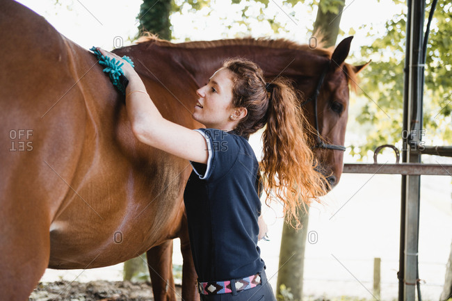 Female equestrian standing in stable and tenderly grooming mane of chestnut horse on ranch