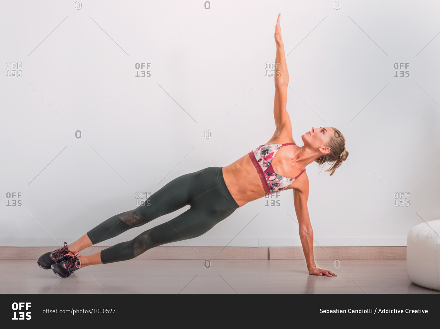 Fit female in sports clothes and sneakers standing in Side Plank pose with arm reached up while practicing yoga in flat near white wall and looking up