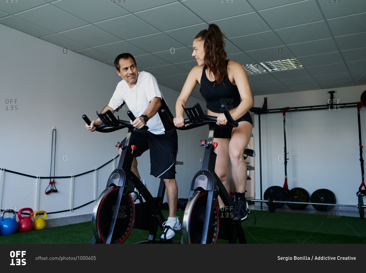Muscular sportsman and sportswoman doing cardio exercises on cycling machines in gym while talking and looking at each other