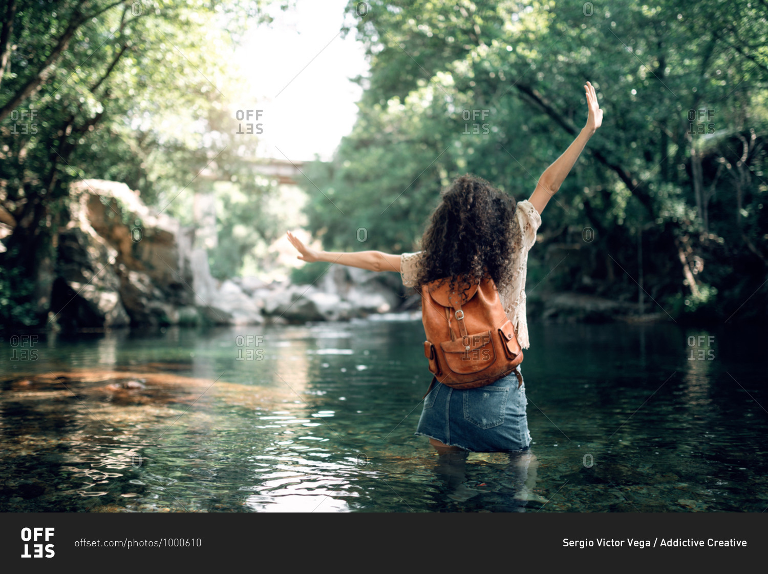 Cheerful female tourist in summer outfit standing in water of river and enjoying vacation with outstretched arms
