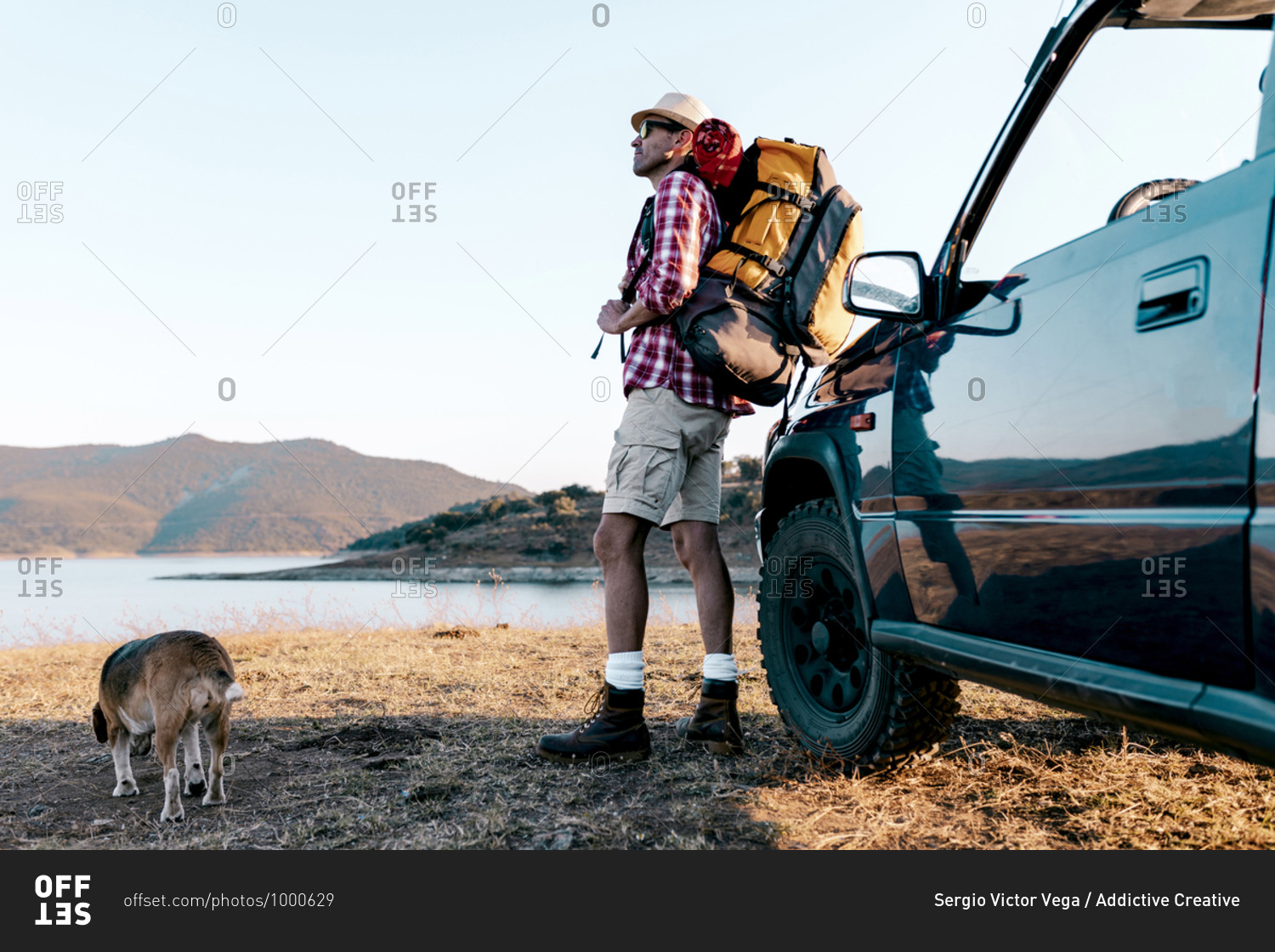 Side view of unrecognizable male traveler in hat with rucksack standing near automobile on faded grass while travelling with dog and contemplating picturesque highland with mountains and river in summer