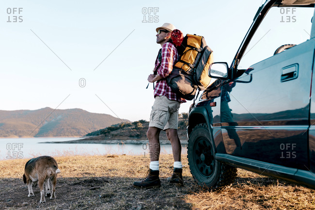 Side view of unrecognizable male traveler in hat with rucksack standing near automobile on faded grass while travelling with dog and contemplating picturesque highland with mountains and river in summer