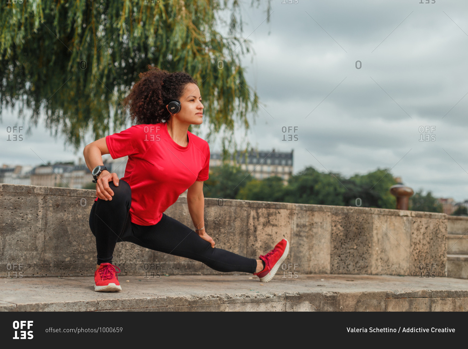 Pensive fit ethnic female athlete in active wear squatting while stretching leg and listening to music in wireless headset on embankment and looking away under cloudy sky