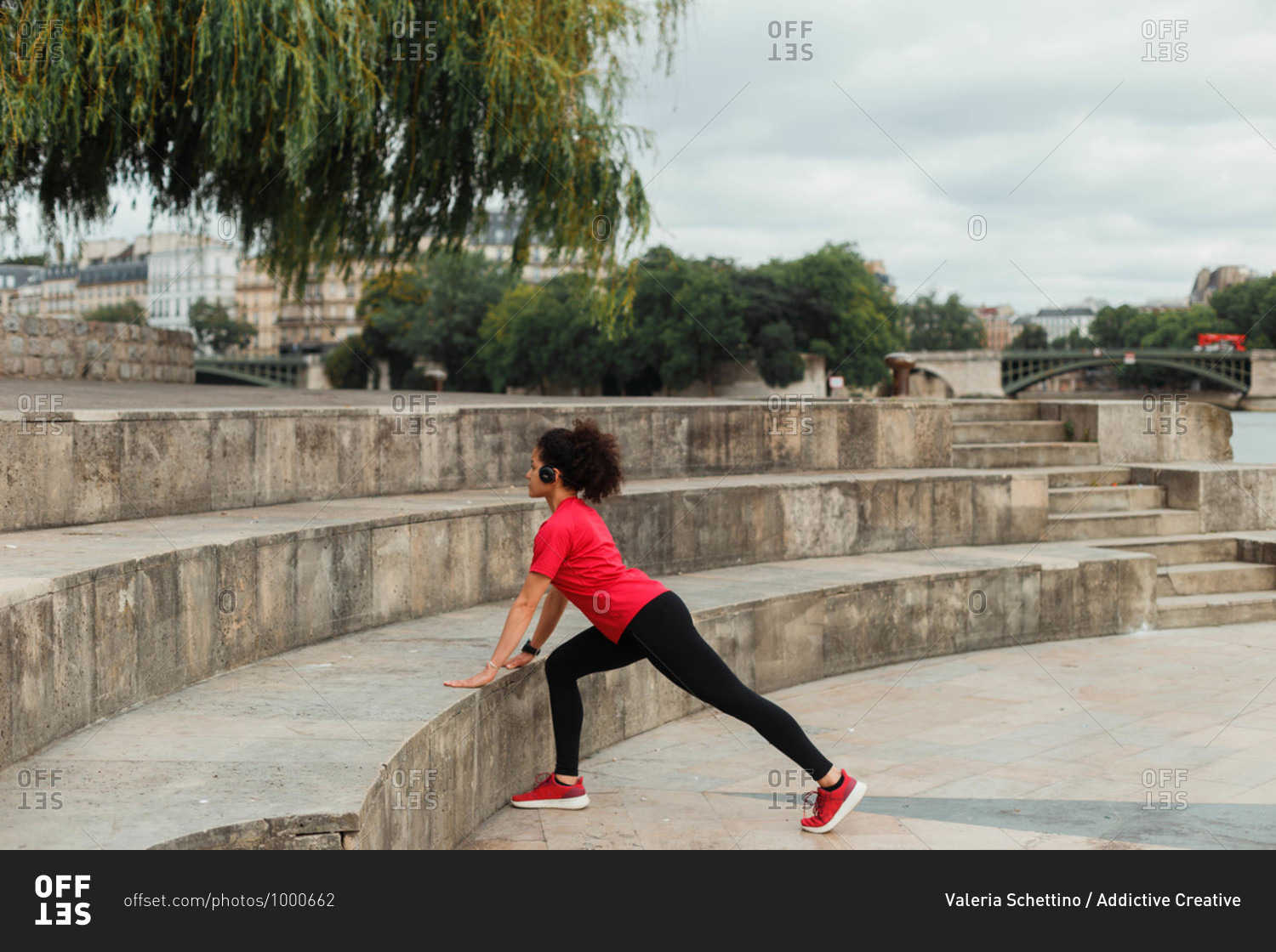 Side view of unrecognizable slim ethnic sportswoman in active wear and headset during training and leaned on staircase near trees in city under cloudy sky
