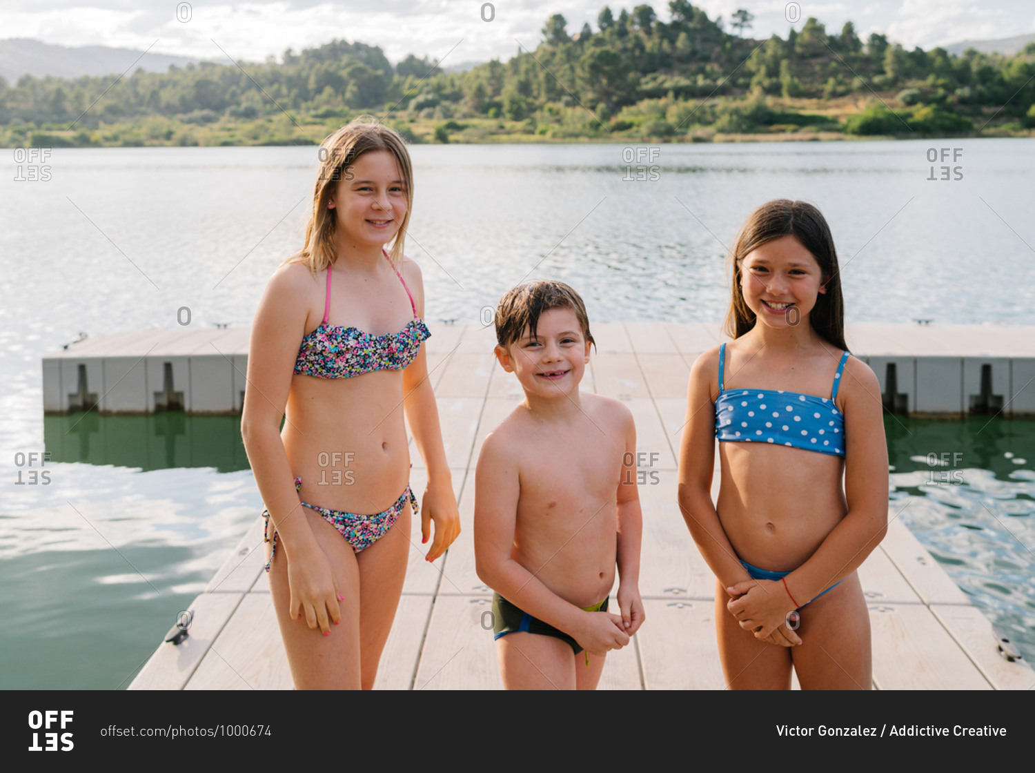 Smiling brother and sisters in swimwear standing on quay near lake and  looking at camera while enjoying summer vacation stock photo - OFFSET