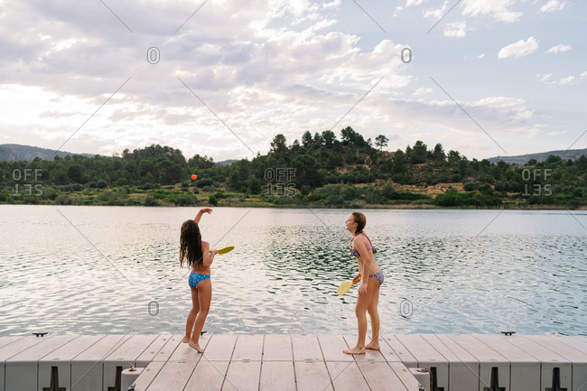 Side view of teenage girls in bikini playing beach tennis with ball and rackets while standing on wooden quay and enjoying summer holiday