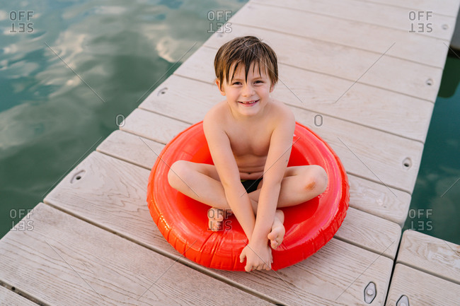 Adorable preteen child with wet hair looking at camera relaxing on inflatable ring on wooden pier during summer vacation near lake