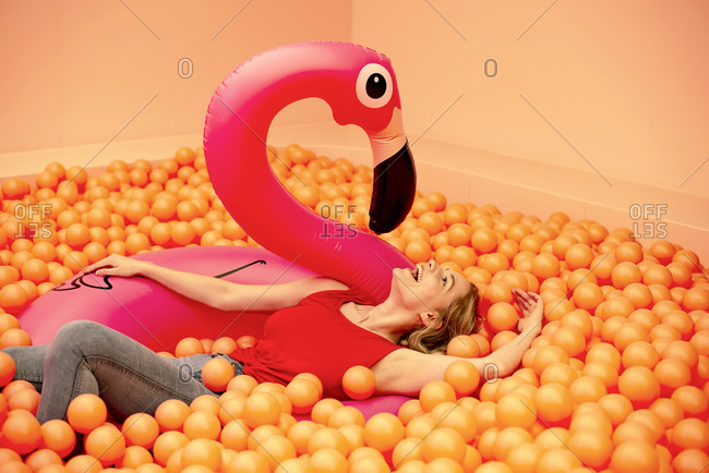 Cheerful young woman with inflatable flamingo lying in orange ball pit