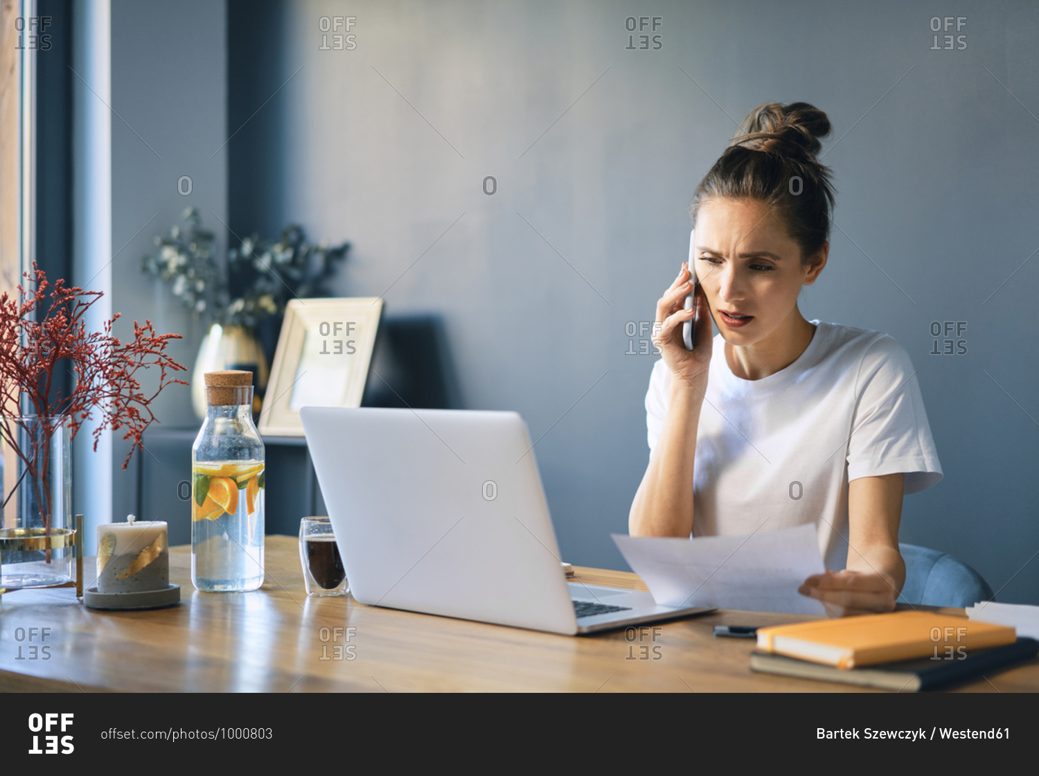 Young woman holding document talking over smart phone at desk in home office