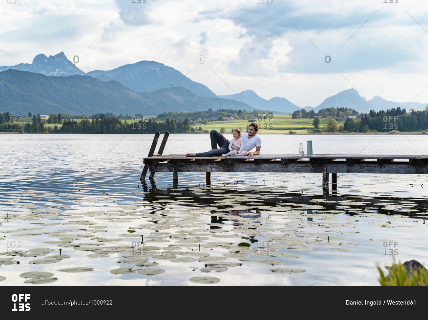 Father and daughter looking at view while relaxing on jetty over lake against mountains