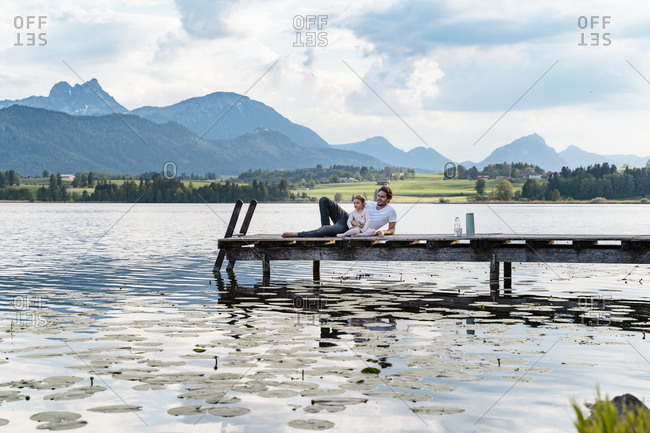 Father and daughter looking at view while relaxing on jetty over lake against mountains