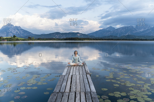 Woman meditating while sitting on jetty over lake against cloudy sky