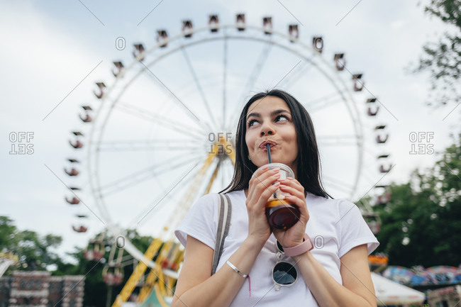 Young woman drinking soft drink while standing against Ferris wheel in amusement park