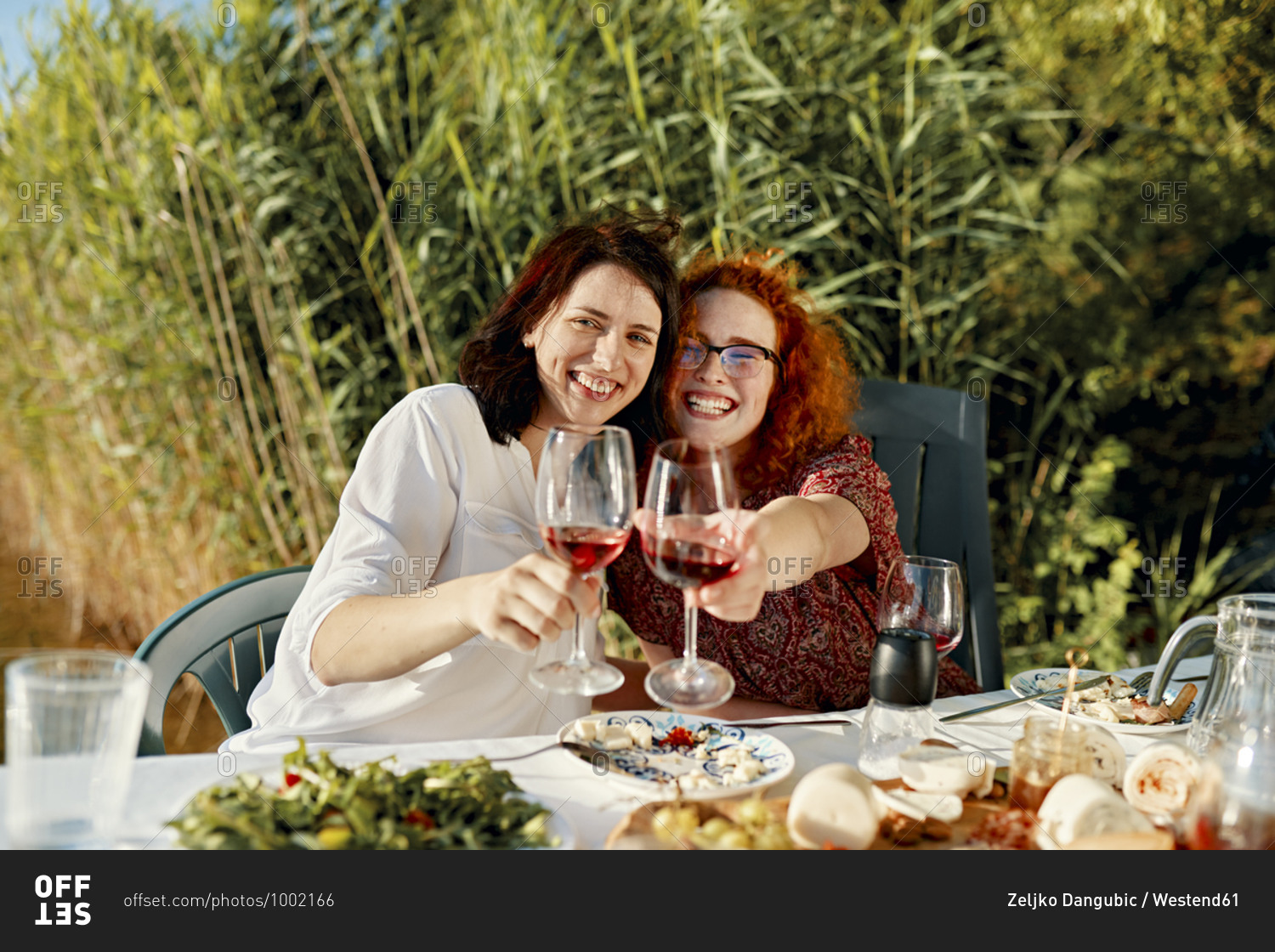Portrait of two happy female friends having dinner at the lakeside raising wine glasses