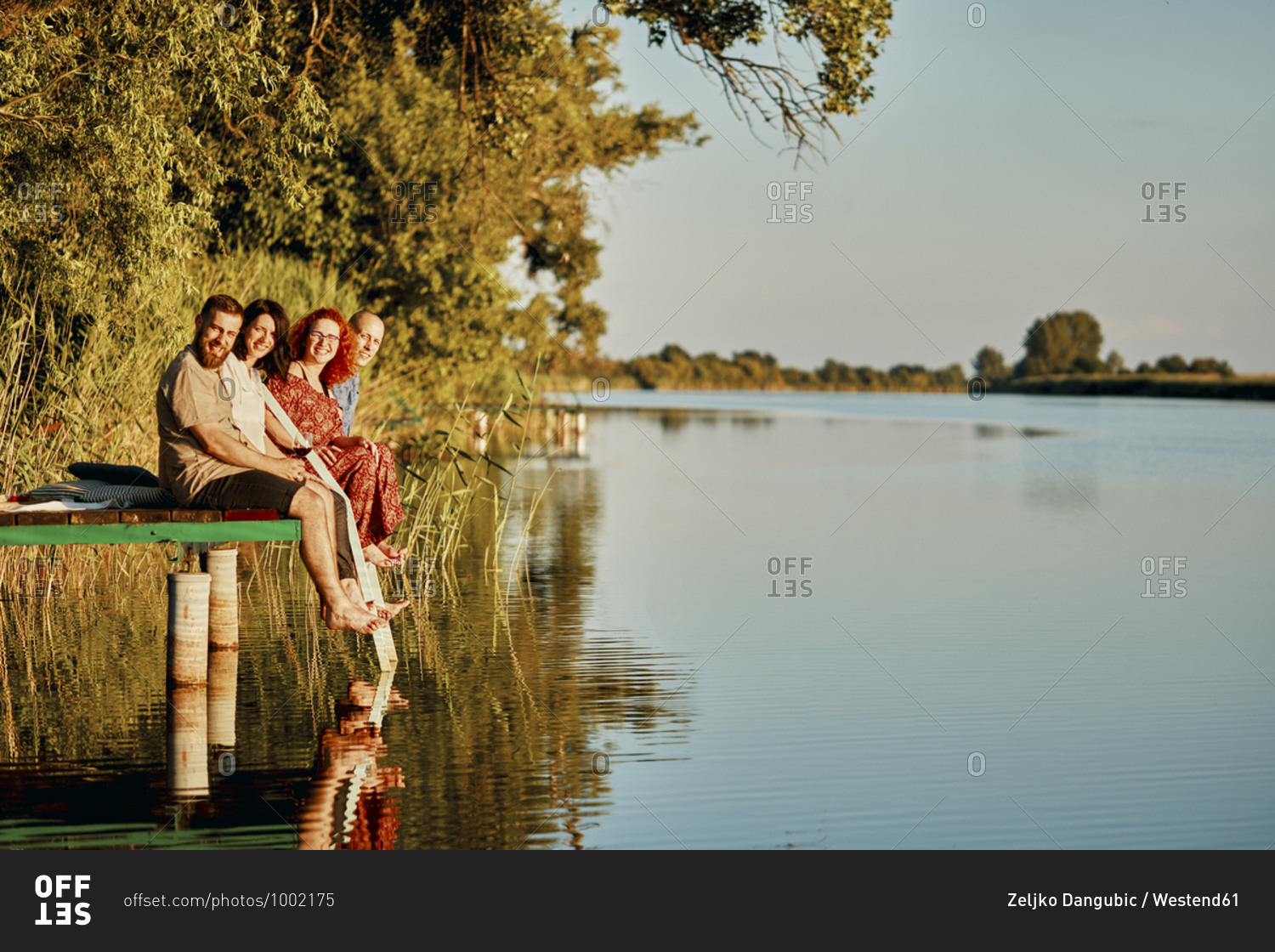 Portrait of smiling friends reflected in water sitting on jetty at a lake