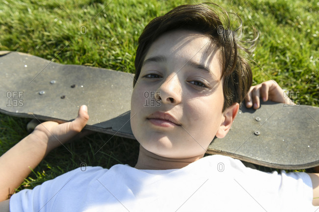 Boy with brown hair lying on grass, head resting on skateboard, looking at camera.