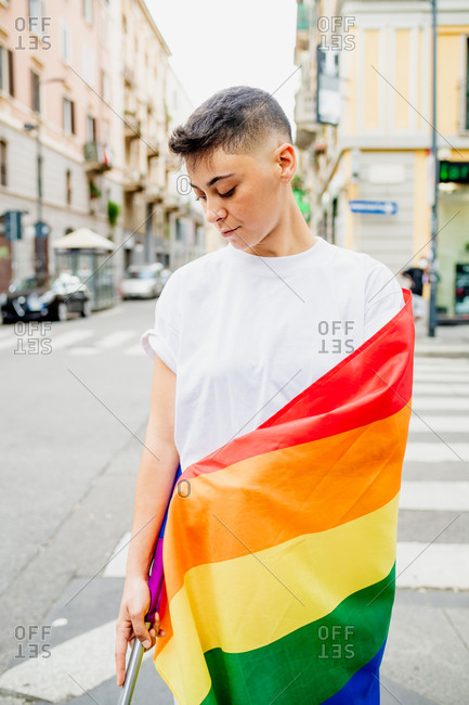 Young lesbian woman standing on a street, wrapped in rainbow flag. stock  photo - OFFSET