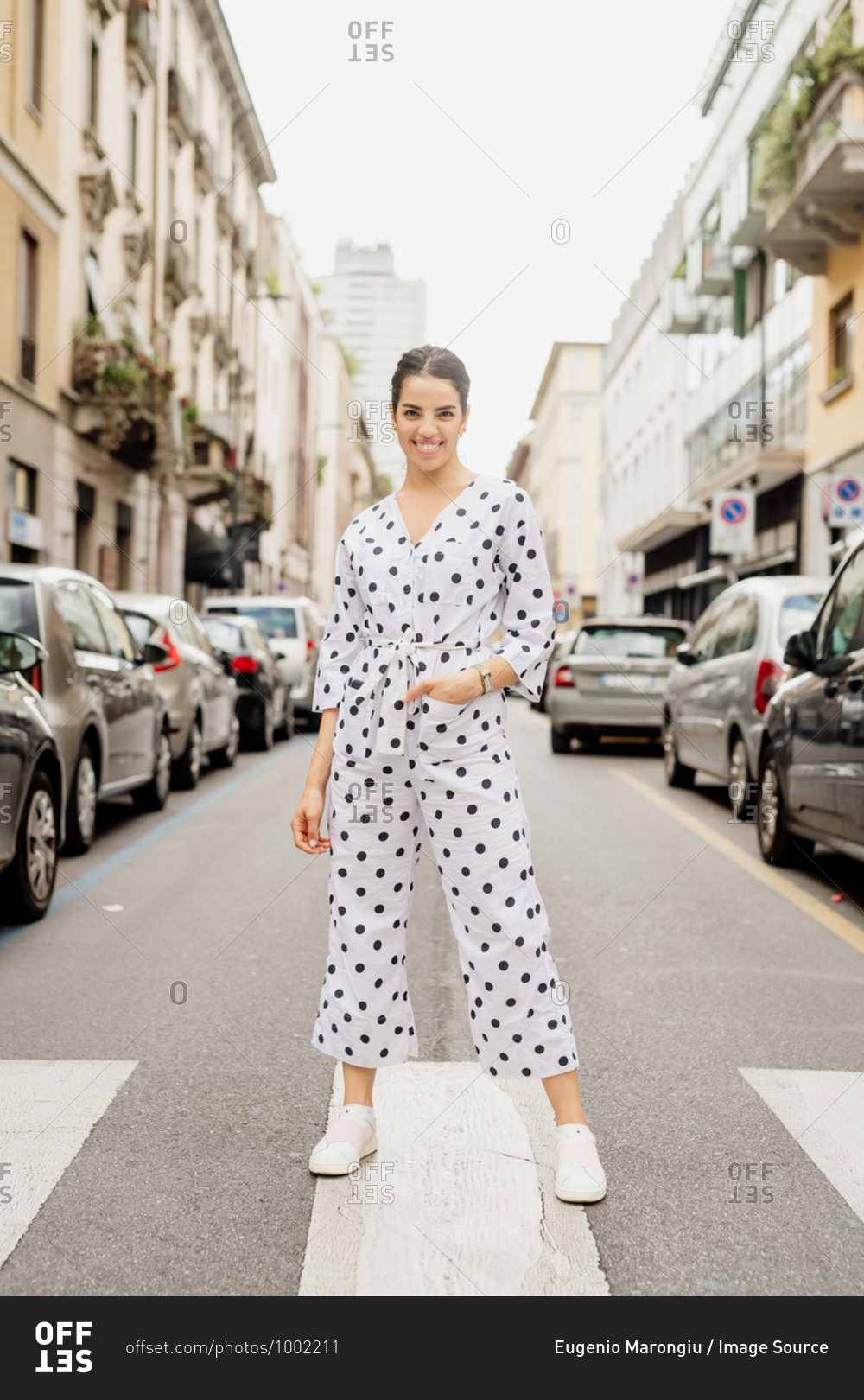 Portrait of smiling woman wearing white and black polka dot jumpsuit, standing on pedestrian crossing.
