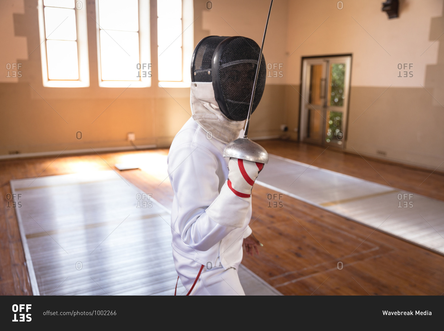 Caucasian sportswoman wearing protective fencing outfit during a fencing training session, preparing for a duel, holding an epee in front of face. Fencers training at a gym.